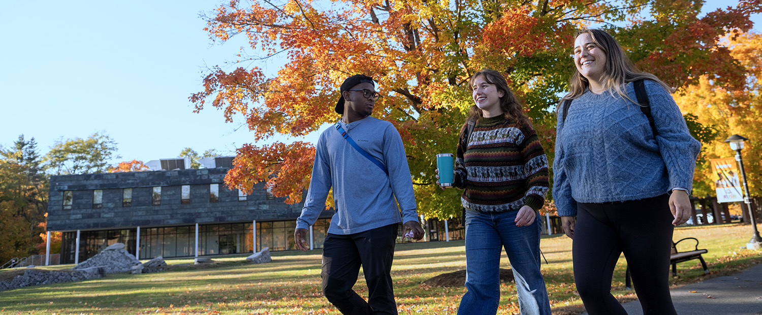 Three students walking together on a campus path in front of autumn trees and a gray building.